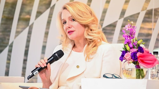 Shell event speaker seated with microphone and tablet, next to a table with flowers and water bottles.